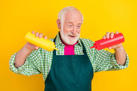 Portrait of attractive trendy elderly cheerful grey-haired man waiter adding sauce making tasty dish isolated over bright yellow color backgroundの写真素材