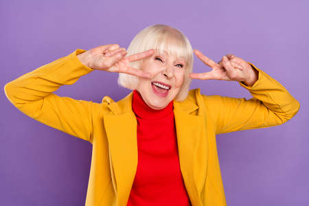 Photo of funny cute short hairdo senior lady show v-sign wear yellow jacket isolated on purple color backgroundの写真素材