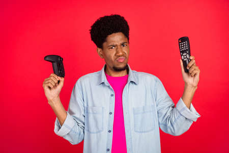 Photo of young afro guy frown hold joystick remote controller choice decision isolated over red...