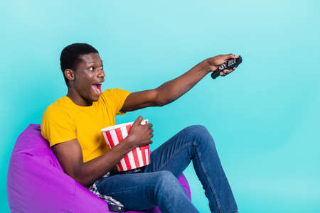 Profile photo of funny brunette hairdo young man eat pop corn watch tv wear yellow t-shirt isolated...