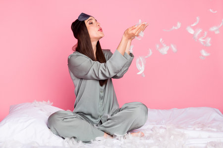 Photo of funny shiny young lady sleepwear sitting blanket blowing feather isolated pastel pink color backgroundの写真素材