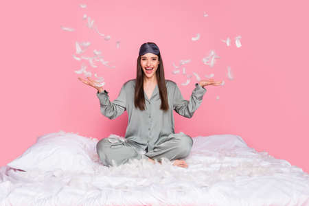 Photo of adorable funny young lady sleepwear smiling sitting blanket throwing feather isolated pastel pink color backgroundの写真素材