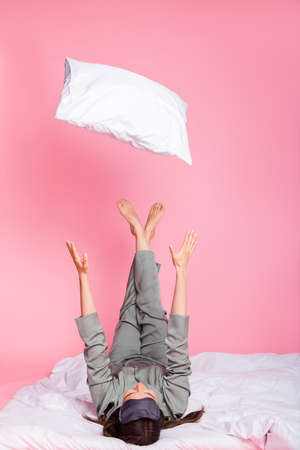 Vertical photo of childish sweet young woman nightwear lying duvet throwing pillow isolated pastel pink color backgroundの写真素材