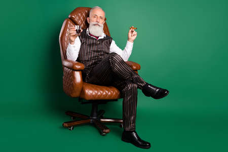Photo of rich english gentleman sit chair drink whiskey smoke cigar wear striped suit on green color...