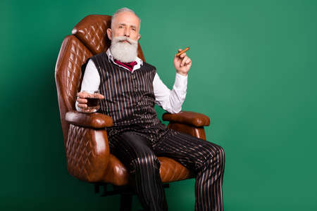 Photo of old noble aristocrat sit armchair drink whiskey enjoy cigar wear striped suit on green color backgroundの写真素材