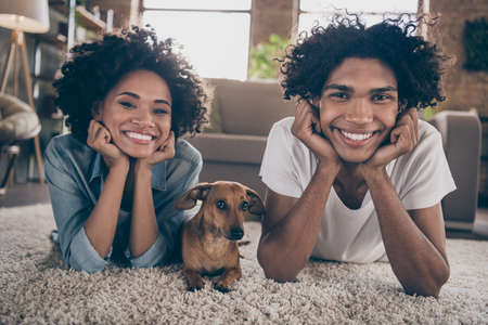 Photo of happy positive dark skin young couple lie carpet dog lover enjoy weekend indoors inside house home flatの写真素材