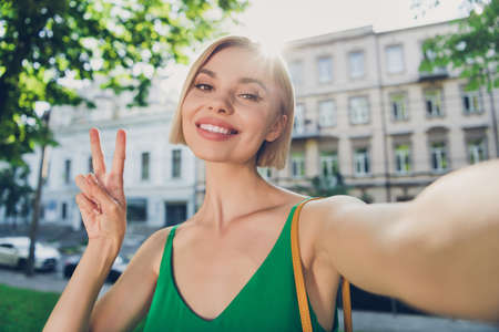 Photo of pretty adorable young lady wear green outfit handbag smiling walking showing v-sign tacking...