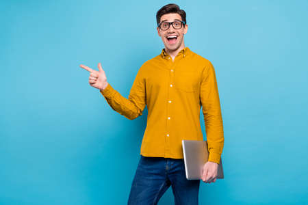 Photo of excited guy hold laptop indicate forefinger empty space wear yellow shirt isolated blue...