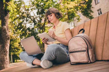 Full body photo of young serious woman write note hold laptop sit bench urban lifestyle outside outdoors city townの写真素材