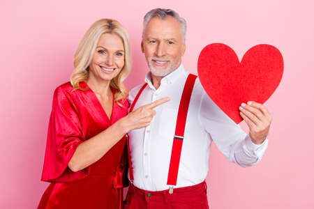 Portrait of two beautiful handsome trendy cheery life partners demonstrating heart shape card isolated over pink pastel color backgroundの写真素材