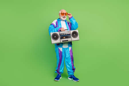Photo of funny pretty senior man wear sportive suit dark eyewear listening boombox smiling isolated green color backgroundの写真素材