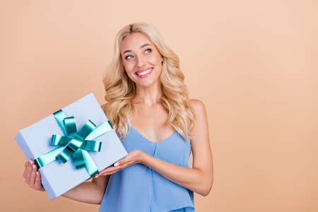Photo of thoughtful sweet young lady wear blue singlet smiling holding gift box looking empty space isolated beige color backgroundの写真素材