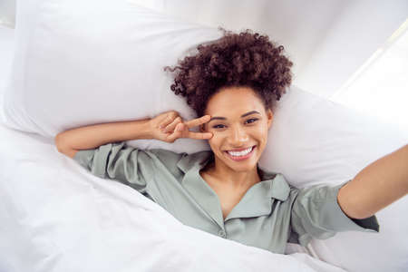 Self-portrait of attractive cheerful funky wavy-haired girl lying in bed showing v-sign at light...