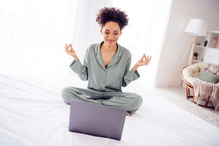 Portrait of attractive dreamy cheerful girl sitting on bed using laptop meditating at light home...