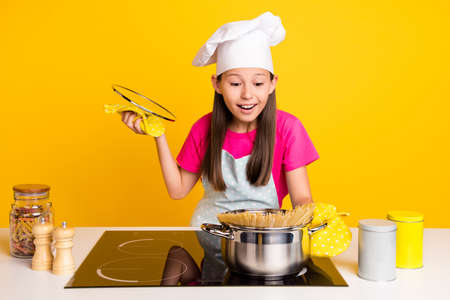 Photo of excited surprised little girl cook pasta check process wear glove apron cap isolated yellow color backgroundの写真素材