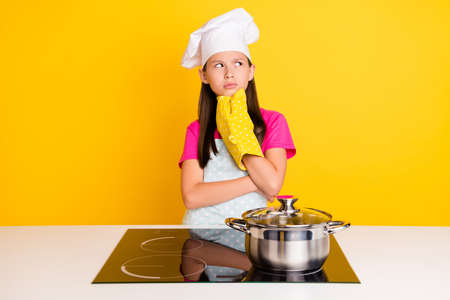 Photo of minded uncertain small girl look empty space table cooker wear glove apron cap isolated yellow color backgroundの写真素材