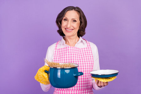 Photo of nice cooker white aged lady hold pan wear white shirt apron isolated on purple color backgroundの写真素材