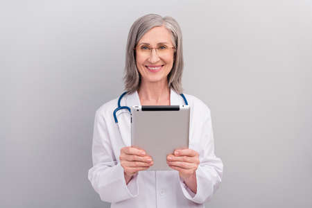 Photo of charming positive mature lady doctor wear white coat spectacles smiling typing modern gadget isolated grey color backgroundの写真素材