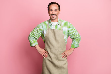 Photo of pretty confident young guy dressed beige apron plaid shirt smiling arms hands waist isolated light pastel pink color backgroundの写真素材