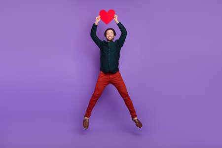 Photo of funny sweet young man wear smart casual outfit smiling rising big red heart jumping high isolated purple color backgroundの写真素材