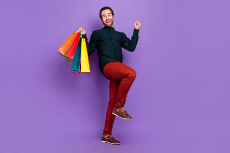 Photo of funny lucky young guy dressed smart casual clothes walking rising fist shopping smiling isolated violet color backgroundの写真素材