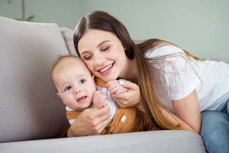 Photo of charming dreamy lady wear white t-shirt smiling sitting sofa hugging baby inside indoors...