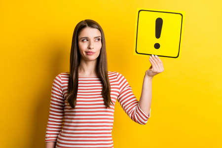 Portrait of attractive doubtful girl holding exclamation point card deciding isolated over bright yellow color backgroundの写真素材