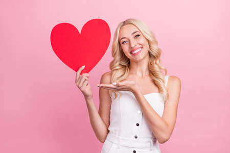 Photo of cute sweet young woman dressed white overall pointing arm big paper heart smiling isolated pink color backgroundの写真素材
