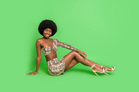 Full length photo of sexy shiny dark skin lady wear print costume sitting floor smiling isolated green color backgroundの写真素材