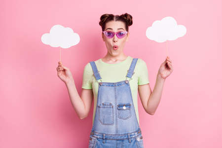 Photo of amazed girl hold two cloud sticks wear sunglass jeans overall green t-shirt isolated on...