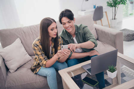 Photo of pretty attractive young lovers dressed casual clothes sitting sofa reading modern device smiling indoors apartmentの写真素材