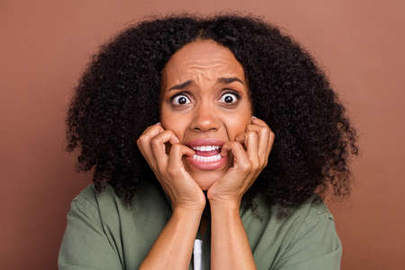 Photo of scared brunette young afro hairstyle lady biting nails wear khaki shirt isolated on brown color backgroundの写真素材