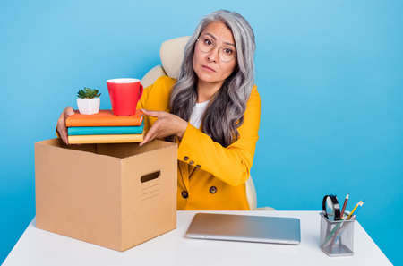 Photo of unhappy depressed mature woman wear yellow jacket spectacles packing stuff lost work isolated blue color backgroundの写真素材