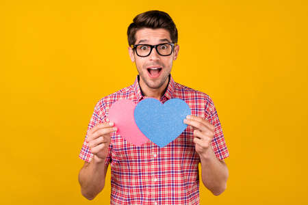 Portrait of attractive amazed cheerful guy holding getting together two heart sign amour isolated over bright yellow color backgroundの写真素材