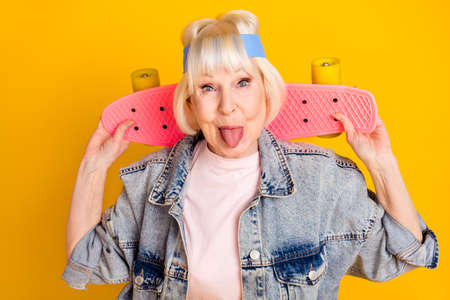 Photo portrait of elder woman keeping skateboard showing tongue feel young isolated on bright yellow color backgroundの写真素材