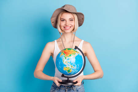 Photo of attractive happy young woman tourist hold hands globe wear hat isolated on blue color backgroundの写真素材
