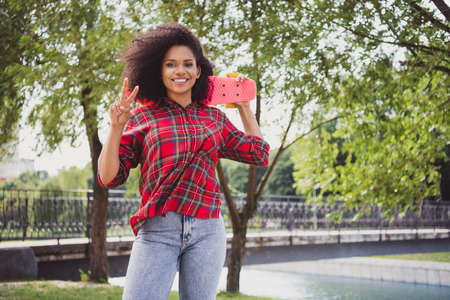 Portrait of attractive cheerful girl wearing checked shirt holding skate showing v-sign on fresh air...