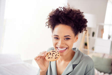 Portrait of attractive cheerful wavy-haired girl sitting in bed eating tasty fresh cookies staying...