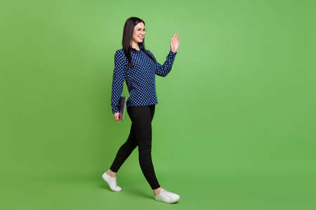 Full length profile photo of business lady go hold laptop wave hand wear spectacles blue shirt trousers isolated on green color backgroundの写真素材