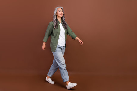 Full length photo of nice elder white hairdo lady go wear khaki shirt jeans sneakers isolated on brown color backgroundの写真素材