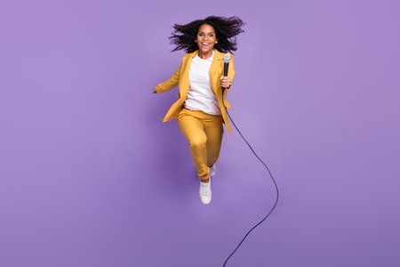 Photo of adorable pretty lady wear yellow suit glasses jumping holding microphone isolated purple color backgroundの写真素材
