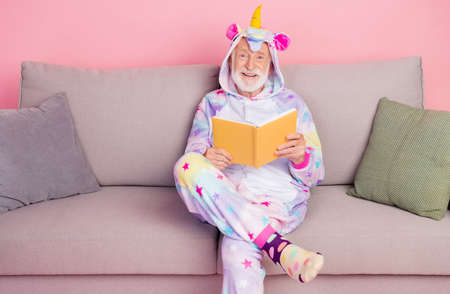 Portrait of attractive cheerful grey-haired man sitting on divan reading novel pastime isolated over pink color backgroundの写真素材