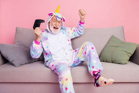 Portrait of handsome trendy cheerful grey-haired man sitting on divan playing game rejoice isolated...