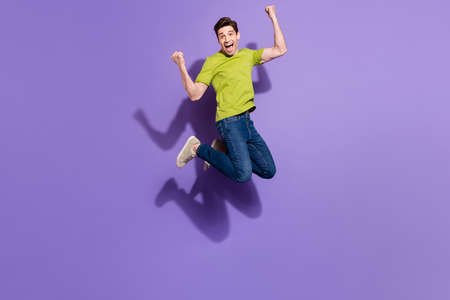 Photo of funny lucky young guy dressed green t-shirt smiling jumping high rising fists isolated...