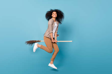 Photo of cute shiny dark skin lady wear plaid shirt smiling jumping riding broom isolated blue color...