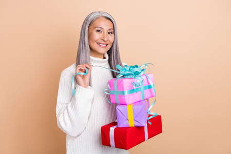 Photo of cheerful funny lady hold stack giftboxes unpack present wear white pullover isolated beige color backgroundの写真素材
