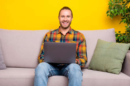 Photo of cheerful man sit couch hold netbook remote job concept wear plaid shirt isolated yellow color backgroundの写真素材