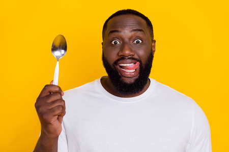 Photo of funny positive handsome guy hold spoon tongue lick teeth lips isolated on yellow color backgroundの写真素材
