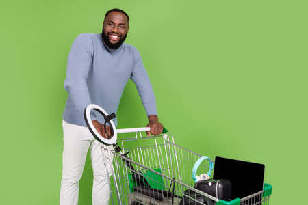 Photo of cheerful african guy dressed sweater walking shopping tray gadgets isolated green color...