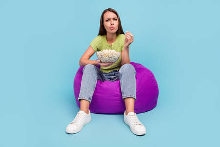 Full body photo of unsure brunette millennial lady sit on bag eat pop corn wear t-shirt jeans sneakers isolated on blue color backgroundの写真素材